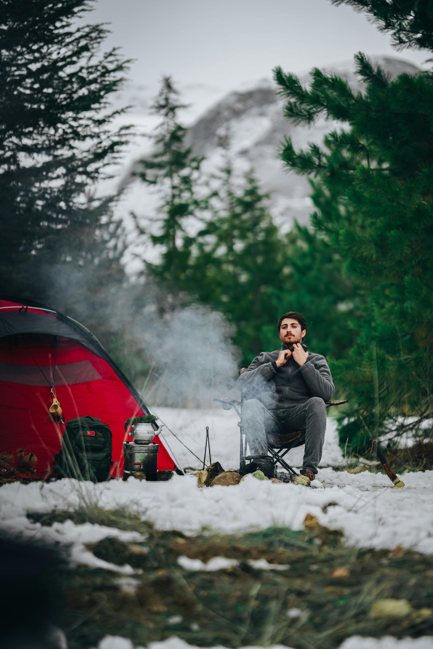 A man sitting by a red tent during a winter camping trip in a snowy forest.
