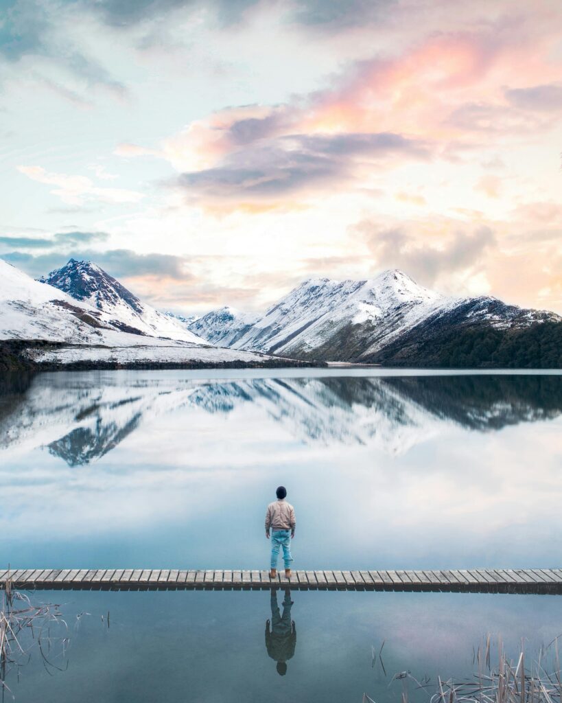 Person standing by a serene lake with snow-capped mountains in Otago, New Zealand.